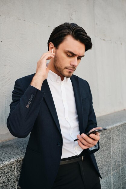 Young beautiful serious bearded brunette man in white shirt and classic suit with wireless earphones thoughtfully using cellphone while standing near gray wall on street