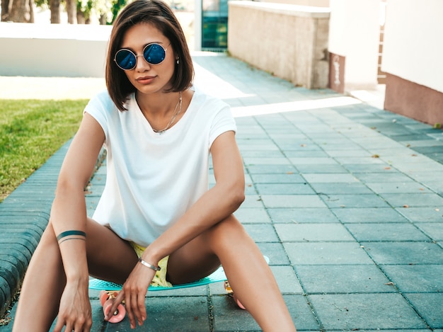 Foto gratuita giovane bella donna sexy hipster fiducioso in occhiali da sole. ragazza di tendenza in t-shirt e pantaloncini estivi. femmina positiva con skateboard penny blu in posa sullo sfondo strada