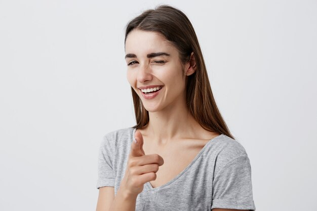 Young beautiful sexy dark-haired caucasian girl with long hair in stylish plain t-shirt smiling with teeth, winking, pointing with finger, posing with happy and satisfied expression.