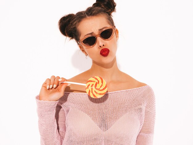 Young beautiful sexy hipster woman with red lips in sunglasses.Trendy girl in summer casual clothes. Positive female going crazy.Funny model isolated on whiteTwo horns.Eating candy lollipop.Makes duck