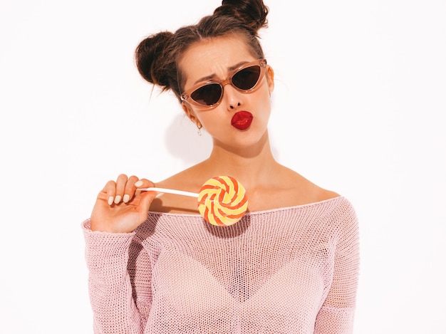 Young beautiful sexy hipster woman with red lips in sunglasses.Trendy girl in summer casual clothes. Positive female going crazy.Funny model isolated on whiteTwo horns.Eating candy lollipop.Makes duck