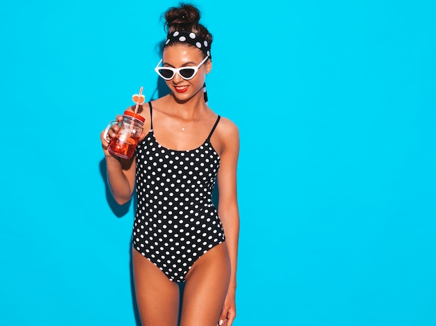 Young beautiful sexy smiling hipster woman in sunglasses.Girl in summer peas swimwear bathing suit.Posing near blue wall,drinking fresh cocktail smoozy drink