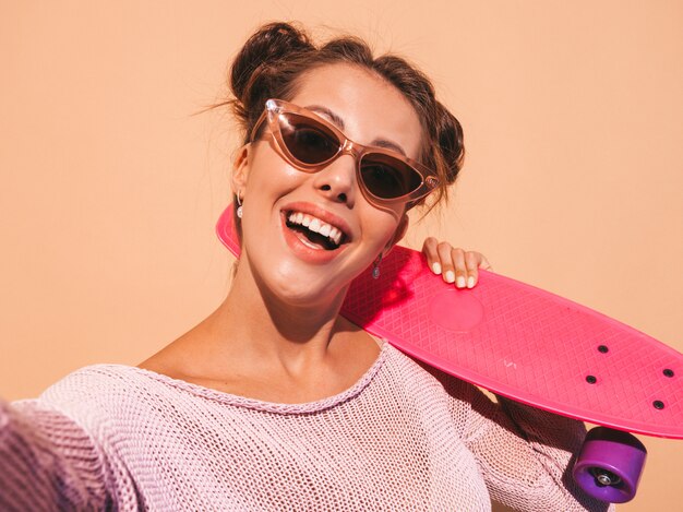 Foto gratuita giovane bella donna sorridente sexy hipster in occhiali da sole. ragazza di tendenza in cardigan lavorato a maglia estiva. femmina con skateboard penny rosa, isolato sulla parete beige. scattare foto di autoritratto selfie su phon