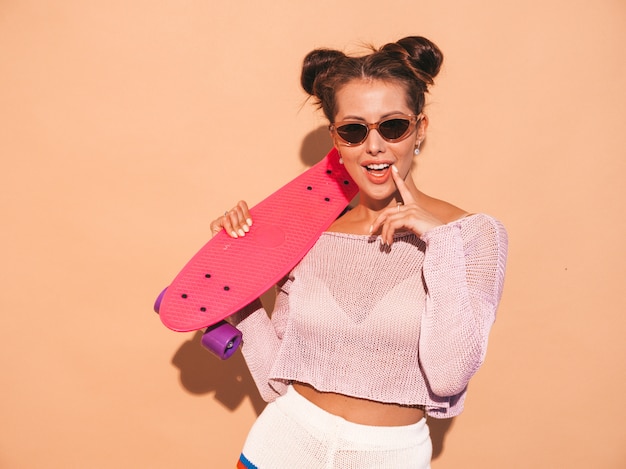 Free photo young beautiful sexy smiling hipster woman in sunglasses.trendy girl in summer knitted cardigan topic ,shorts.positive female going crazy with pink penny skateboard,isolated on beige wall.two hor