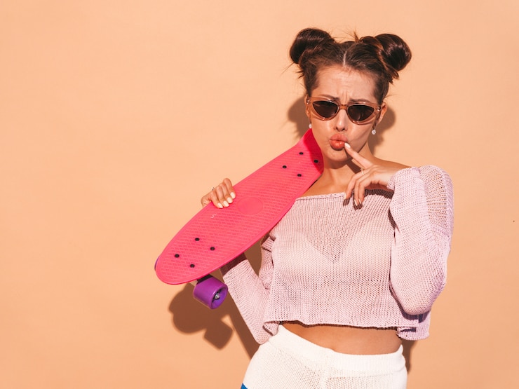 Free Photo young beautiful sexy smiling hipster woman in sunglasses.trendy girl in summer knitted cardigan topic ,shorts.positive female going crazy with pink penny skateboard,isolated on beige wall.two hor