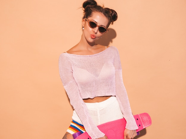 Free photo young beautiful sexy smiling hipster woman in sunglasses.trendy girl in summer knitted cardigan topic ,shorts.positive female going crazy with pink penny skateboard,isolated on beige wall.two hor