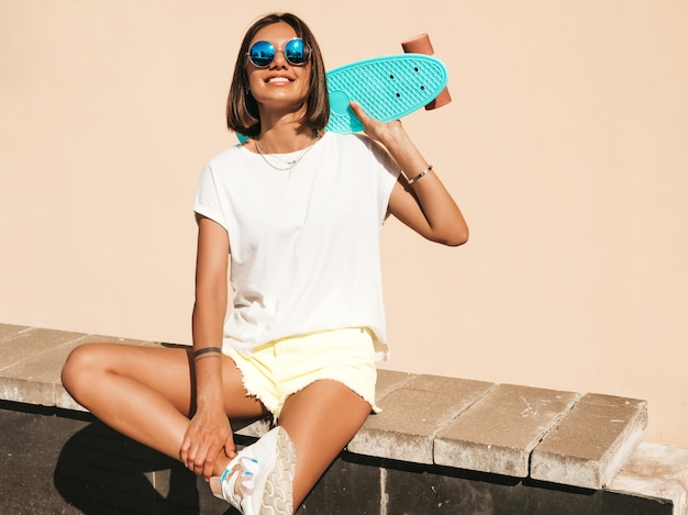 Young beautiful sexy smiling hipster woman in sunglasses.Trendy girl in summer T-shirt and shorts.Positive female with blue penny skateboard posing on the street background