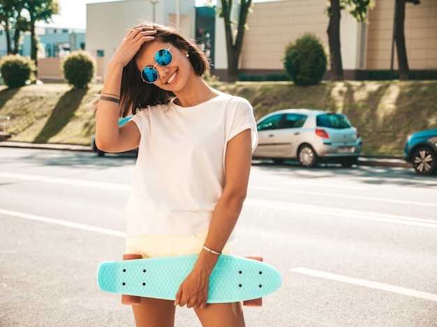 Giovane bella donna sorridente sexy dei pantaloni a vita bassa in occhiali da sole ragazza alla moda in maglietta e shorts di estate femmina positiva con il pattino blu del penny che posa sui precedenti della via