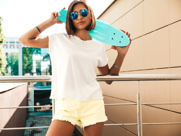 Young beautiful sexy smiling hipster woman in sunglasses.Trendy girl in summer T-shirt and shorts.Positive female with blue penny skateboard posing on the street background