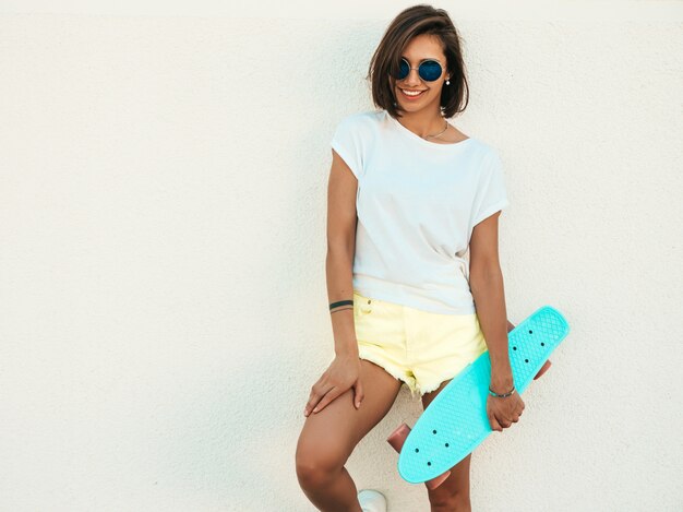 Молодая красивая сексуальная улыбающаяся хипстерская женщина в солнцезащитных очках. Модная девушка в летней футболке и шортах. Позитивная самка с синим пенни скейтбордом позирует на улице возле белой стены