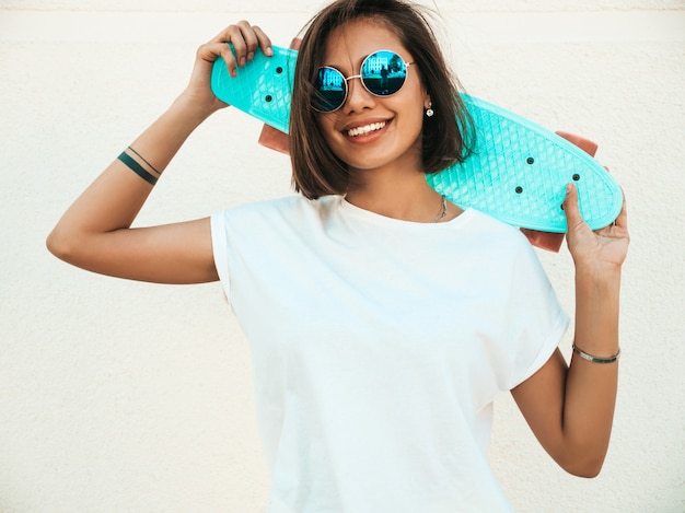 Foto gratuita giovane bella donna sorridente sexy dei pantaloni a vita bassa in occhiali da sole. ragazza alla moda in maglietta e shorts di estate. femmina positiva con il pattino blu del penny che posa nella via vicino al muro bianco