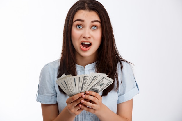 Young beautiful shoked business girl holding money over white wall