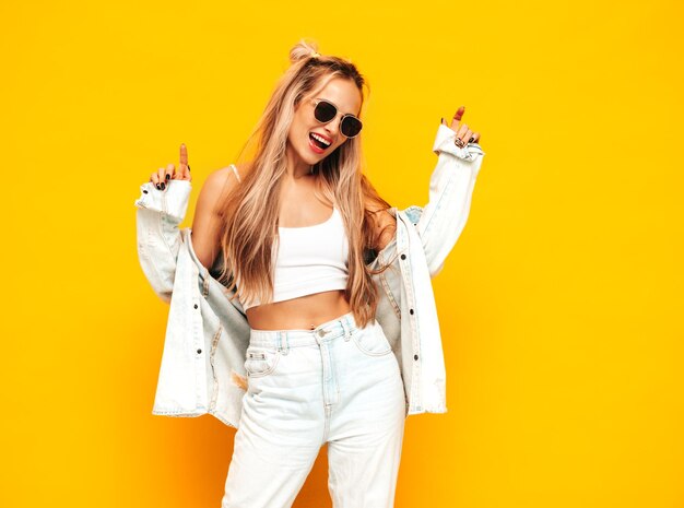 Young beautiful smiling blond female in trendy summer clothes Sexy carefree woman posing near yellow wall in studio Positive model having fun indoors Cheerful and happy in sunglasses Dancing