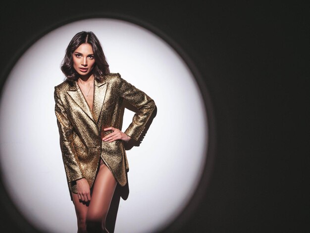 Young beautiful smiling brunette female in trendy evening gold jacket Sexy carefree woman posing near white wall in studio in a circle of lightFashionable model with bright makeup