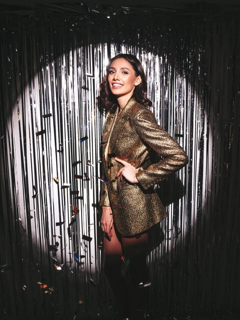 Young beautiful smiling brunette female in trendy evening golden jacket Sexy carefree woman posing near silver shiny tinsel wall in studioFashionable model with bright makeup looks at camera