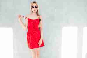 Бесплатное фото Молодая красивая улыбающаяся женщина в модном летнем красном платье сексуальная беззаботная блондинка позирует у стены в студии позитивная модель веселится веселая и счастливая в солнечный день