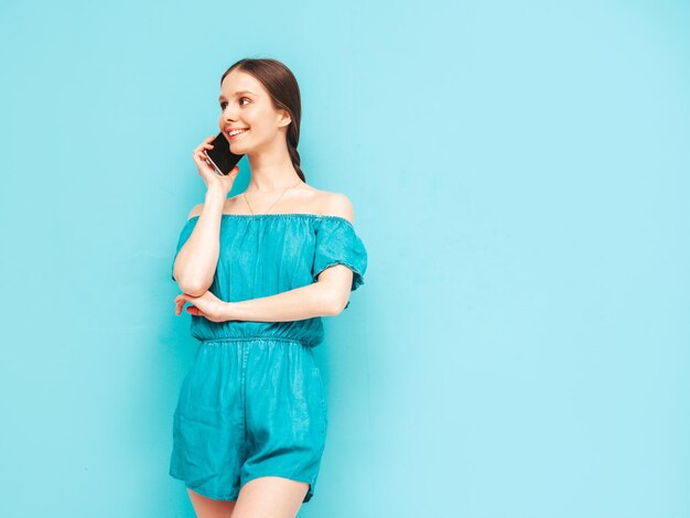 トレンディな夏の青いオーバーオールで若い美しい笑顔の女性スタジオで壁の近くでポーズをとるセクシーな屈託のない女性携帯電話を使用してポジティブブルネットモデルスマートフォンで話す