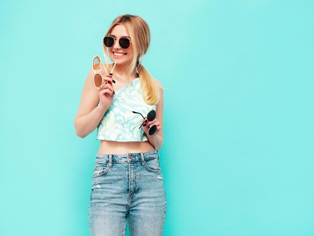 Young beautiful smiling female in trendy summer clothes Sexy carefree woman posing near blue wall in studio Positive model holding many sunglassesCheerful and happy Showing and making choice