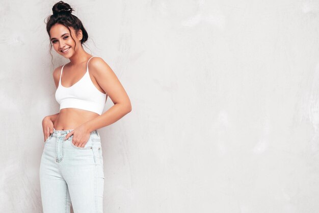 Young beautiful smiling female in trendy summer clothes Sexy carefree woman posing near grey wall in studio Positive brunette model having fun and going crazy Cheerful and happy