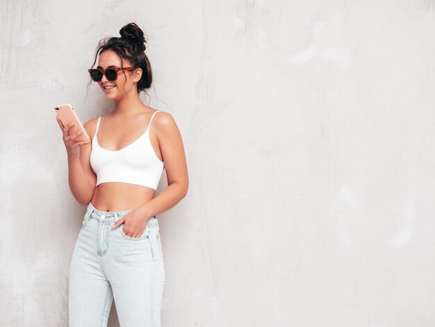 Young beautiful smiling female in trendy summer clothes Sexy carefree woman posing near grey wall in studio Positive brunette model having fun and going crazy Using smartphone apps Holding phone