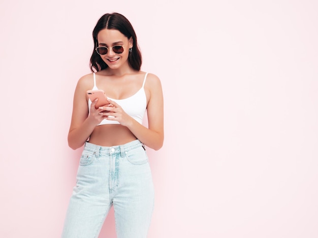 Молодая красивая улыбающаяся женщина в модной летней одежде Сексуальная беззаботная женщина позирует возле розовой стены в студии Позитивная брюнетка модель смотрит на экран мобильного телефона Держа смартфон и используя приложения