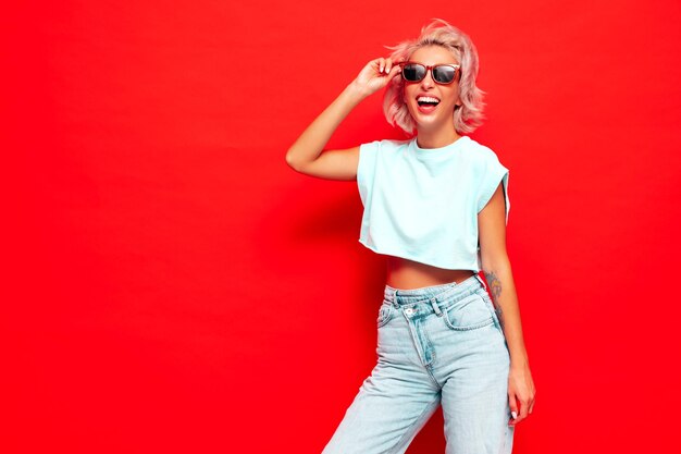 Young beautiful smiling female in trendy summer clothes Sexy carefree woman posing near red wall in studio Positive blond model having fun and going crazy Cheerful and happy In sunglasses