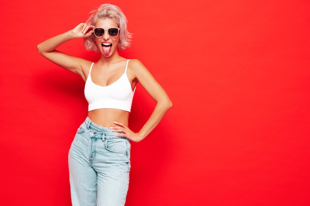 Young beautiful smiling female in trendy summer clothes Sexy carefree woman posing near red wall in studio Positive blond model shows tongue and going crazy Cheerful and happy In sunglasses