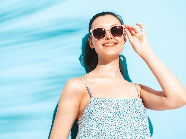Молодая красивая улыбающаяся женщина в модном летнем платье Сексуальная беззаботная женщина позирует возле синей стены в студии Позитивная брюнетка модель веселится Веселая и счастливая в солнечных очках Изолированные