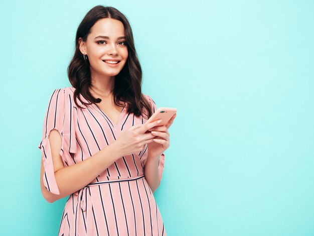 Young beautiful smiling female in trendy summer dress Sexy carefree woman posing near blue wall in studio Positive brunette model looking at cellphone screen Holding smartphone and using apps