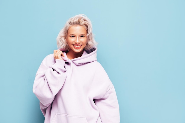 Young beautiful smiling female in trendy summer hoodie Sexy carefree woman posing near blue wall in studio Positive blond model having fun and going crazy Cheerful and happy