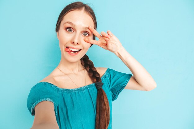 Young beautiful smiling female in trendy summer jeans overalls Sexy carefree woman with tail hairstyle posing near wall in studio Positive model having fun Cheerful and happy Taking selfie