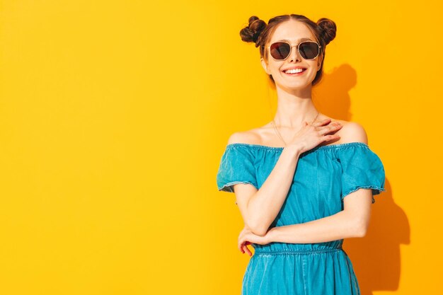 Young beautiful smiling female in trendy summer jeans overalls Sexy carefree woman with two horns hairstyle posing near yellow wall in studio Positive model having fun Isolated In sunglasses