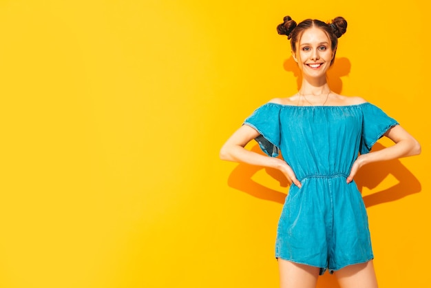 Young beautiful smiling female in trendy summer jeans overalls Sexy carefree woman with two horns hairstyle posing near yellow wall in studio Positive model having fun Isolated