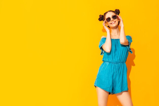 Young beautiful smiling female in trendy summer jeans overalls Sexy carefree woman with two horns hairstyle posing near yellow wall in studio Positive model having fun Isolated