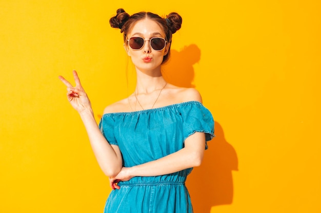 Young beautiful smiling female in trendy summer jeans overalls sexy woman with two horns hairstyle posing near yellow wall in studio positive model having fun shows peace sign in sunglasses