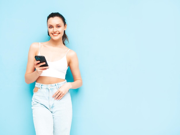 최신 유행의 여름 청바지를 입은 젊고 아름다운 웃는 여성 스튜디오의 파란색 벽 근처에서 포즈를 취하는 섹시한 평온한 여성 스마트폰을 들고 앱을 사용하여 휴대폰 화면을 보고 있는 긍정적인 갈색 머리 모델