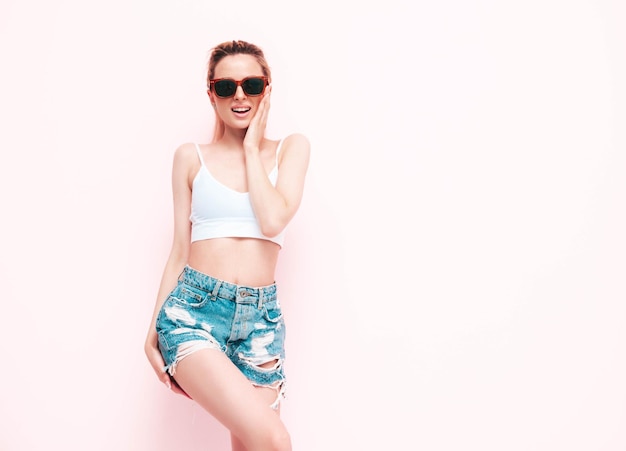 Young beautiful smiling female in trendy summer jeans shorts Sexy carefree blond woman posing near wall in studio Positive model having fun Cheerful and happy in sunglasses Isolated
