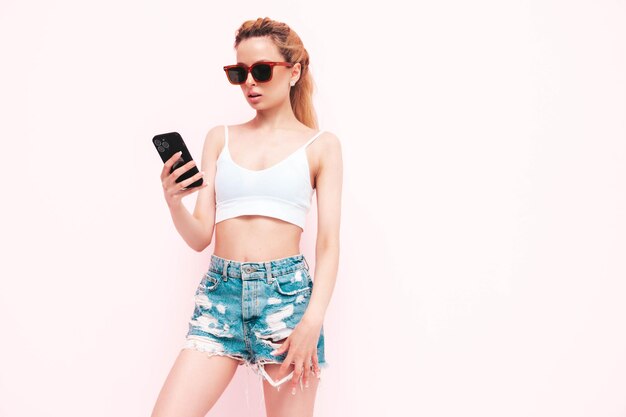 Молодая красивая улыбающаяся женщина в модных летних джинсовых шортах Сексуальная беззаботная блондинка позирует у стены в студии Позитивная модель веселится, держа смартфон и используя приложения для мобильного телефона