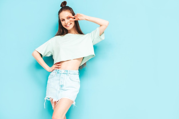Young beautiful smiling female in trendy summer jeans skirt carefree woman posing near blue wall in studio Positive model having fun indoors Cheerful and happy