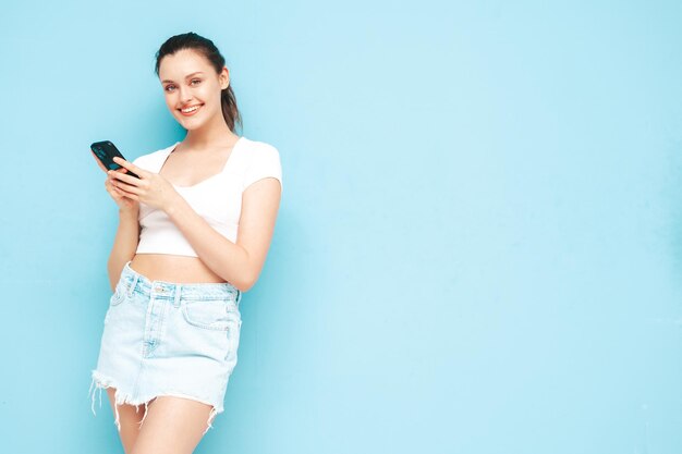 Young beautiful smiling female in trendy summer jeans skirt Sexy carefree woman posing near blue wall in studio Positive brunette model looking at cellphone screen Holding smartphone and using apps