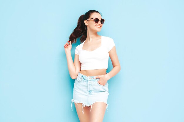 Молодая красивая улыбающаяся женщина в модной летней джинсовой юбке и верхней одежде Сексуальная беззаботная женщина позирует возле синей стены в студии Позитивная брюнетка-модель веселится