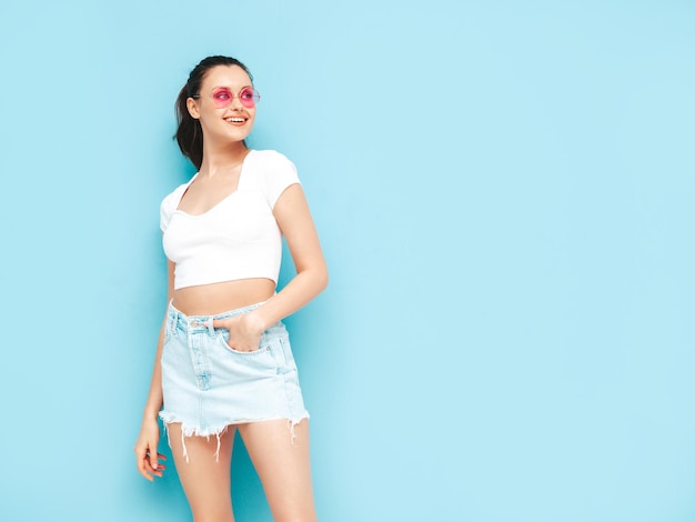 Young beautiful smiling female in trendy summer jeans skirt and top clothes Sexy carefree woman posing near blue wall in studio Positive brunette model having funCheerful and happy