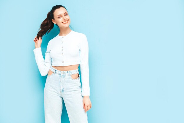 Young beautiful smiling female in trendy summer jeans and top clothes Sexy carefree woman posing near blue wall in studio Positive brunette model having funCheerful and happy