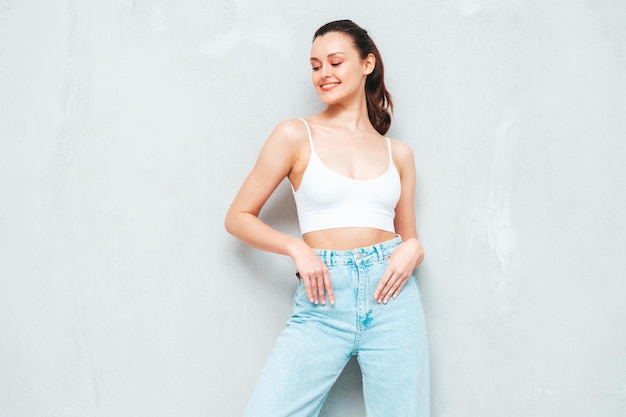 Young beautiful smiling female in trendy summer jeans and top Sexy carefree woman posing near grey wall in studio Positive brunette model having fun and going crazy Cheerful and happy