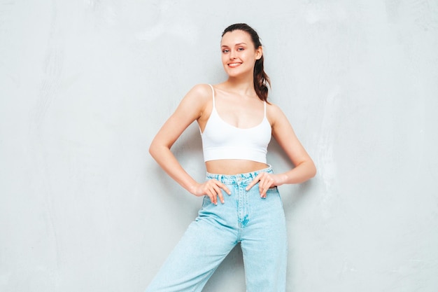 Free photo young beautiful smiling female in trendy summer jeans and top sexy carefree woman posing near grey wall in studio positive brunette model having fun and going crazy cheerful and happy