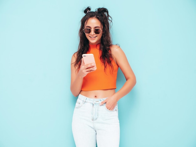 Молодая красивая улыбающаяся женщина в модном летнем оранжевом топе и джинсовой одежде Сексуальная беззаботная женщина позирует у синей стены в студии Глядя на экран смартфона Модель с помощью приложений для мобильного телефона