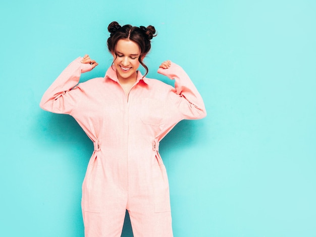 Free photo young beautiful smiling female in trendy summer pink overalls sexy carefree woman posing near blue wall in studio positive model having fun and going crazy cheerful and happy dancing at party