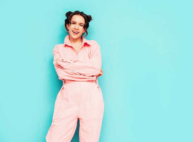 Young beautiful smiling female in trendy summer pink overalls Sexy carefree woman posing near blue wall in studio with two horns Positive model having fun and going crazy Cheerful and happy