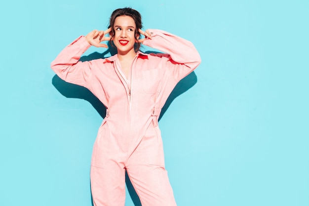 Young beautiful smiling female in trendy summer pink overalls Sexy carefree woman posing near blue wall in studio with two horns Positive model having fun and going crazy Cheerful and happy