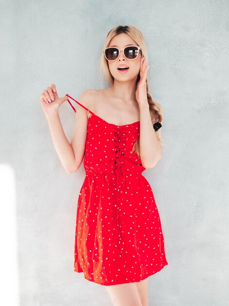 トレンディな夏の赤いドレスで若い美しい笑顔の女性スタジオの壁の近くでポーズをとるセクシーな屈託のないブロンドの女性サングラスで陽気で幸せな楽しみを持っているポジティブモデル